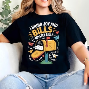 May include: Black t-shirt with a graphic of a yellow mailbox with the text "I bring joy and bills - mostly bills." The design includes paper airplanes and hearts. The shirt is made of a soft material.