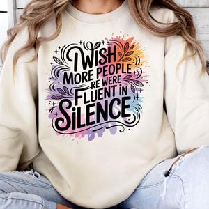 May include: Cream-colored sweatshirt with the text "Wish More People Were Fluent In Silence" in a black, stylized font. The text is surrounded by colorful watercolor-style splashes of pink, purple, orange, and blue.