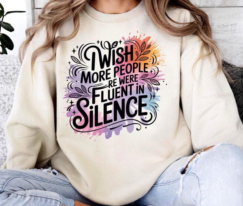 May include: Cream-colored sweatshirt with the text "Wish More People Were Fluent In Silence" in a black, stylized font. The text is surrounded by colorful watercolor-style splashes of pink, purple, orange, and blue.