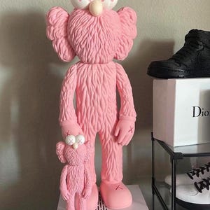 Può includere: Una grande statuetta KAWS BFF rosa in piedi accanto a una statuetta KAWS BFF rosa più piccola abbinata. Entrambe le figure presentano superfici testurizzate simili a pelliccia, teste rotonde con occhi a forma di X e orecchie arrotondate.