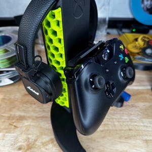 Op de afbeelding: Zwarte Xbox-controller en Marshall-koptelefoon op een zwart-neon groene gaming headset- en controllerstandaard. De standaard heeft een honingraatontwerp en het Xbox-logo.