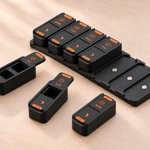 May include: A black weekly pill organizer with seven individual modular containers. Each container features orange text for the days of the week, sun icons for morning, and moon icons for evening. The base tray includes magnetic slots.