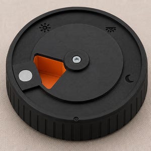Peut inclure: Un récipient circulaire noir avec un couvercle rotatif doté d'une vis argentée au centre. Le couvercle présente des icônes de soleil, de coucher de soleil et de lune. Une ouverture triangulaire révèle un intérieur orange vif et un aimant.
