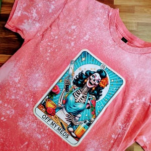 May include: A pink bleached t-shirt with a graphic of a skeleton woman holding a knife and a cup. The graphic is in a card-like frame with the text "OFF MY MEDS" at the bottom.