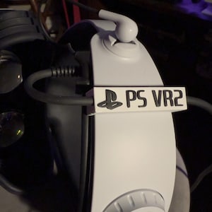 May include: White PlayStation VR2 headset with a black PlayStation logo and the text "PS VR2" on the side. The headset has a white dial on the side and a black cable.