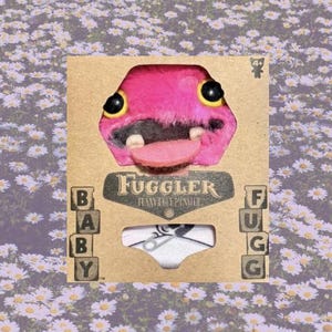 Op de afbeelding: Een roze pluche monster speelgoed met een grote glimlach en een lange tong. Het speelgoed is verpakt in een kartonnen doos met de woorden "Fuggler" en "Furry Ugly Plush" erop gedrukt. De doos bevat ook een klein beeld van een baby die een luier draagt met een veiligheidsspeld.