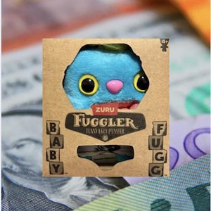 May include: A blue and pink plush toy with large eyes and a pink nose. The toy is in a cardboard box with the words "ZURU FUGGLER FUNNY EGGY MONSTER" printed on it.