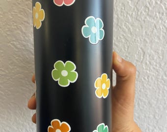 Puede incluir: Botella de agua aislada negra adornada con pegatinas florales de colores. Las pegatinas presentan varios colores, incluyendo rosa, amarillo, azul, verde y naranja, con contornos blancos. La botella es sostenida por una persona.