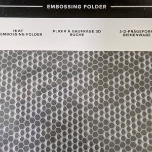 May include: A close-up of a gray and white honeycomb embossing folder. The folder is designed to create a 3D honeycomb effect on paper or cardstock. The text "Hive Embossing Folder" is printed on the folder.