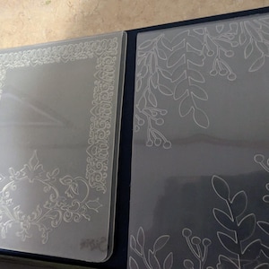 May include: Two clear plastic stencils with intricate floral designs. One stencil features a border design with a floral motif in the corner. The other stencil features a floral design with leaves and berries.