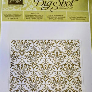 May include: A white card with a gold design of a repeating pattern of swirls and lines. The card has the text "Big Shot" in black and the text "Stampin' Up!" in smaller black text. The card also has the text "Texture Impressions" in black text.