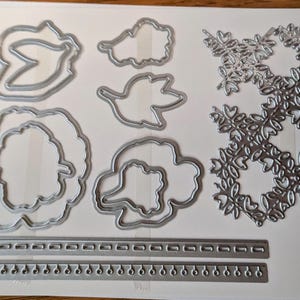 May include: A collection of silver metal die cuts in various floral and leaf shapes, including a large rose, smaller flowers, and decorative borders. The die cuts are arranged on a white surface.
