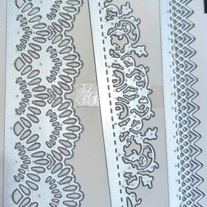 May include: Three silver metal die-cut stencils with intricate designs. The first stencil features a repeating pattern of circles and ovals. The second stencil has a floral design with a repeating pattern of leaves and swirls. The third stencil has a repeating pattern of zigzags.