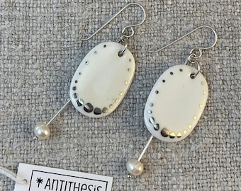May include: A pair of white ceramic earrings with silver accents and a pearl dangle. The earrings are oval shaped and have a silver wire that goes through the top of the earring. The pearl dangle is attached to the bottom of the wire.