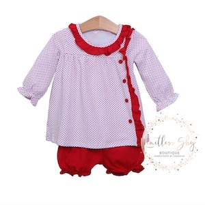 May include: A two-piece baby outfit featuring a white top with red polka dots and ruffled red trim. The top has long sleeves and red buttons. Matching red bloomers complete the set. The outfit is displayed on a wooden hanger.