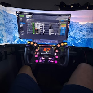 May include: A black sim racing steering wheel with a central digital display showing the number 97 and the name Jackson, surrounded by illuminated pink buttons and rotary dials, positioned in front of a curved monitor displaying a racing interface.