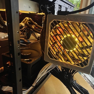May include: A partially assembled black computer case with a power supply unit (PSU) and fan. The PSU is silver with the text "FICKERTEC" and a yellow-lit fan. Cables are neatly arranged.