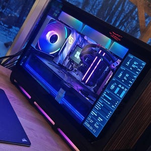 Peut inclure: Un boîtier d'ordinateur personnalisé avec des composants internes éclairés par des LED bleues, violettes et arc-en-ciel. Le boîtier dispose d'un écran numérique affichant des informations système. Un tapis de souris noir est visible au premier plan.