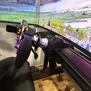 May include: A black racing steering wheel controller with purple accents and multiple buttons, mounted on a black base. The steering wheel is positioned in front of two large computer monitors displaying a landscape image with a taskbar.