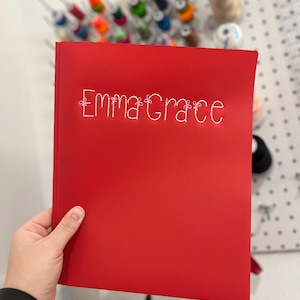 May include: A red binder with the name "Emma Grace" embroidered in white thread. The binder is held in a hand, with a black watch visible on the wrist. Sewing supplies are in the background.