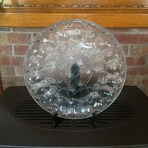 May include: A clear glass decorative plate featuring a textured relief design of pine branches and pinecones. The circular plate has a scalloped edge and is supported by a small black plastic stand, displaying a detailed botanical motif.