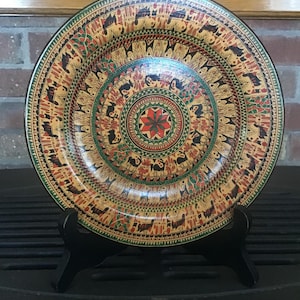 May include: A decorative plate with a detailed, colorful design. The plate features a central floral pattern surrounded by concentric rings of stylized figures in red, green, and black against a tan background. The plate is displayed on a black stand.