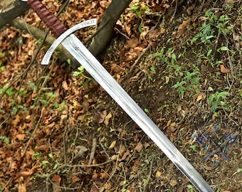 May include: A long, silver sword with a brown leather-wrapped handle and a crossguard. The blade is angled, and the words "CHRISTUS" and "VINCIT" are etched on the crossguard. The sword is resting on a bed of leaves and twigs.