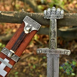 May include: A detailed image of a Viking sword with a patterned blade and ornate silver-colored handle. The handle features intricate knotwork and a crown-like pommel. The wooden scabbard is visible, with silver accents and a deer antler grip.
