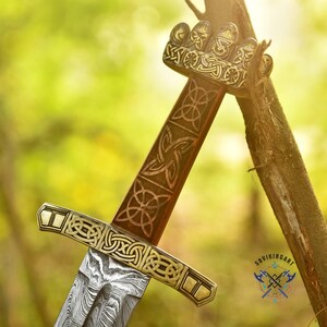 May include: A detailed close-up of a Viking sword. The hilt and guard are adorned with intricate Celtic knotwork and gold accents. The blade features a distinctive pattern. The sword is set against a blurred green background, suggesting an outdoor setting.
