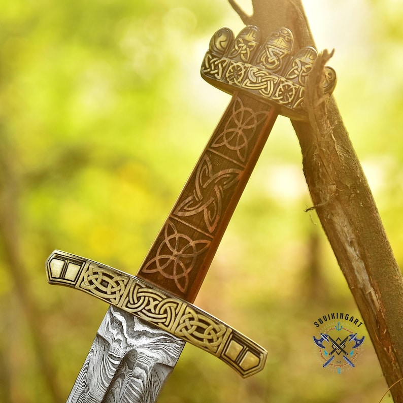 May include: A detailed close-up of a Viking sword. The hilt and guard are adorned with intricate Celtic knotwork and gold accents. The blade features a distinctive pattern. The sword is set against a blurred green background, suggesting an outdoor setting.
