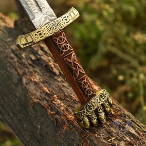 May include: A decorative sword with a patterned blade and a wooden handle. The handle features Celtic knot designs and brass accents. The sword is resting on a weathered log. The blade is approximately 30 cm long.