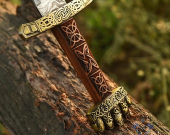 May include: A close-up of a decorative sword with a patterned blade and a wooden handle. The handle features intricate Celtic knot designs and brass accents. The sword is resting on a weathered log.