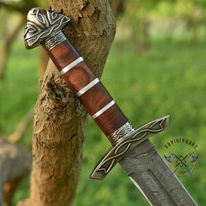 May include: A close-up of a Viking sword with a wooden handle and a silver-colored guard and pommel. The handle is a dark brown color with silver bands. The blade has a patterned design. The sword is leaning against a tree branch.