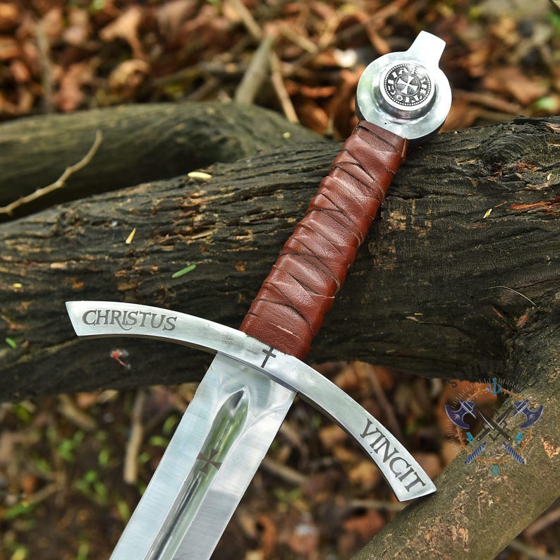 May include: A medieval sword with a silver blade and a brown leather-wrapped handle. The crossguard is inscribed with "CHRISTUS VINCIT". The pommel features a circular design. The sword rests on a wooden log.