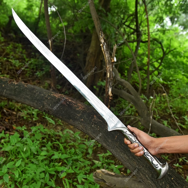 May include: A long, silver sword with a curved blade and ornate handle. The blade has a textured pattern, and the handle features a detailed design. The sword is held against a backdrop of green foliage and a fallen tree.