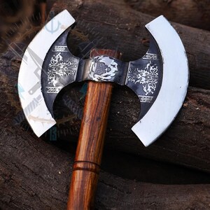 May include: A double-bladed axe with a wooden handle and ornate metalwork. The axe heads are silver, and the central metal piece features engraved designs. The handle is a dark, polished wood.