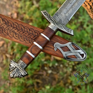 May include: A detailed view of a Viking sword with a patterned blade and a wooden handle with silver accents. The sword rests in a wooden sheath with an engraved design. The handle has a dark brown color, and the sheath is a lighter brown.