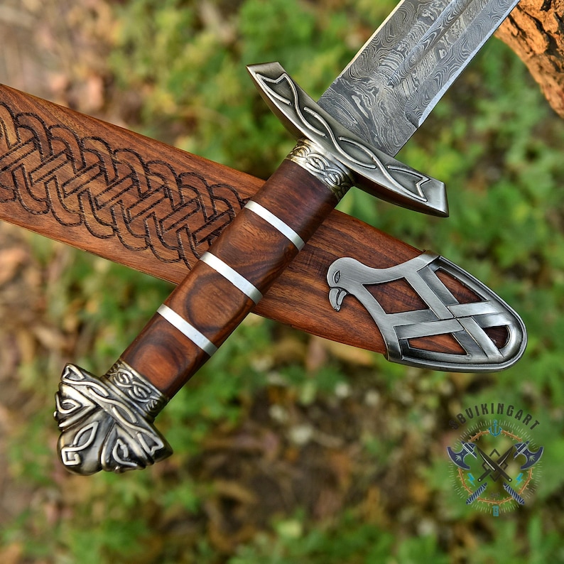 May include: A detailed view of a Viking sword with a patterned blade and a wooden handle with silver accents. The sword rests in a wooden sheath with an engraved design. The handle has a dark brown color, and the sheath is a lighter brown.