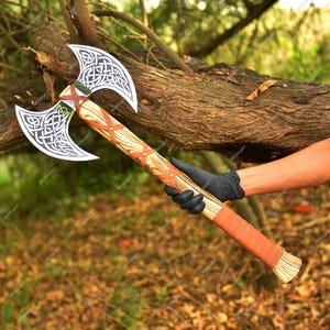 May include: Double-bladed axe with a wooden handle and silver blades. The blades feature intricate Celtic knot designs. The handle is wrapped with brown leather and has a reddish-brown leather cross design. The axe is held by a person wearing a black glove.
