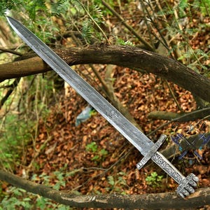 May include: A long, silver sword with a detailed blade pattern and ornate handle. The handle features a hand-shaped grip and intricate carvings. The sword is displayed against a backdrop of natural foliage.