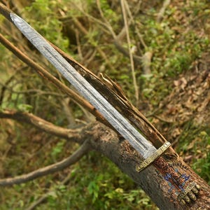 May include: A detailed image of a sword with a patterned blade and ornate handle. The blade has a textured, silver appearance. The handle features gold and brown accents with intricate designs. The sword rests on a tree branch.