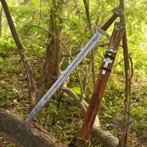 May include: A long, silver sword with a patterned blade and ornate hilt rests against a wooden scabbard. The scabbard is brown with decorative carvings and a white cross. The sword and scabbard are set against a backdrop of trees and foliage.
