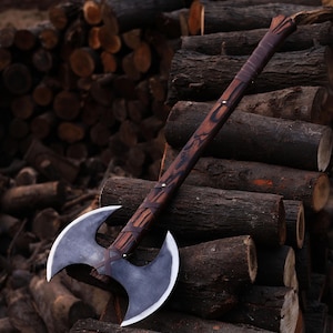 Puede incluir: Un hacha de doble filo con un mango de madera marrón oscuro y una hoja de metal negro. El hacha está apoyada en una pila de troncos apilados.