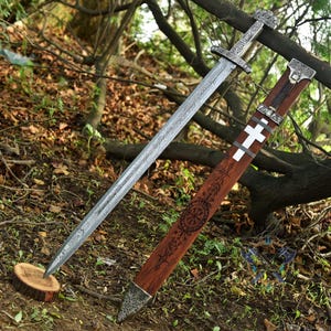 May include: A silver and steel sword with a patterned blade and ornate hilt rests beside its wooden sheath. The sheath is brown with a white cross and etched designs. The sword is displayed outdoors, leaning against a tree branch.
