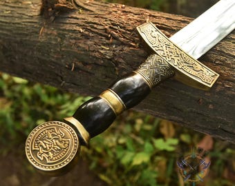 May include: A detailed view of a sword with a silver blade and an ornate handle. The handle features a dark, possibly ebony, grip with gold-colored accents and intricate patterns. The pommel and guard are also gold-colored, with detailed Celtic knot designs.