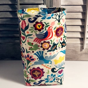 Op de afbeelding: Een witte stoffen opbergbak met een kleurrijk bloemen- en vogelpatroon. Het patroon bevat rode, blauwe, groene, gele en paarse bloemen en vogels.
