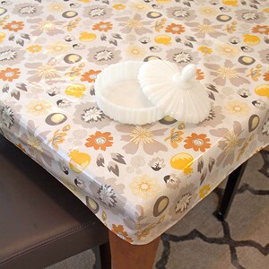 Puede incluir: Un mantel de mesa blanco con flores con un patr&oacute;n floral amarillo y naranja. Un plato de cer&aacute;mica blanco con tapa est&aacute; sobre el mantel.