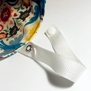 Op de afbeelding: Een witte stoffen band met twee witte plastic drukknopen en een zilveren metalen ring. De band is bevestigd aan een kleurrijke stoffen tas met een bloemenpatroon.
