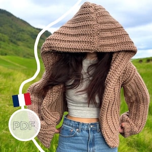 Peut inclure: Un cardigan à capuche en crochet marron avec une texture côtelée. Le cardigan est ouvert sur le devant et a des manches longues. Le modèle porte le cardigan sur un débardeur gris et un jean bleu.