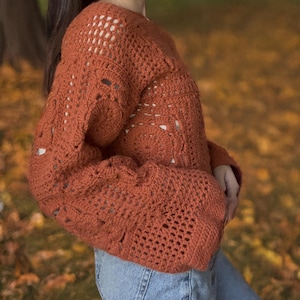 Könnte beinhalten: Ein rostiger, gehäkelter Pullover mit lockerer Passform und Glockenärmeln. Der Pullover ist mit einem zarten, offenen Stichmuster gearbeitet.